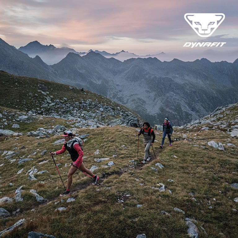 Drei Personen wandern mit Ausrüstung auf einem schmalen Bergpfad durch eine felsige Hochgebirgslandschaft. Rechts oben im Bild ist das DYNAFIT‑Logo zu sehen.