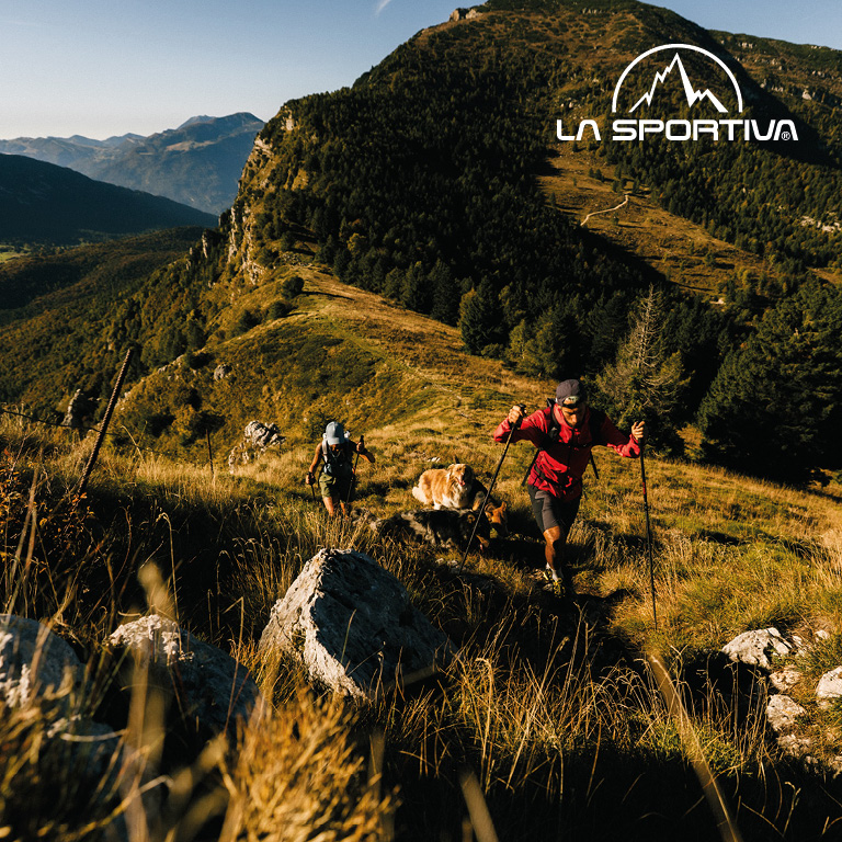 Zwei Personen wandern mit Stöcken einen sonnigen Berghang hinauf, umgeben von Gras und Felsen. Rechts oben im Bild steht das LA SPORTIVA‑Logo.
