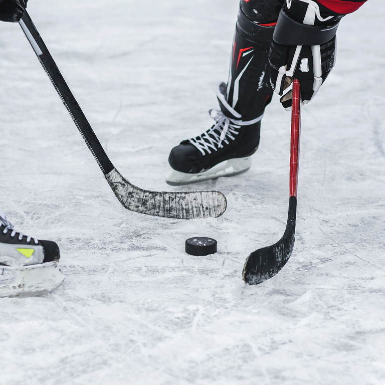 Zu sehen sind zwei SPieler mit Eislaufschuhen und Eishockeyschläger die sich den Puck spielen