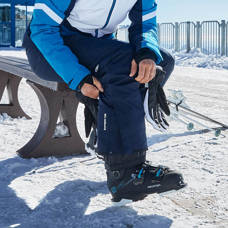 Ein Mann der auf einer Bank sitzt im Schnee und sich seine Hose bei den Skischuhe richtet