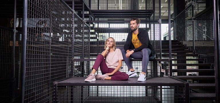 Zwei Personen sitzen auf einer Metallplattform innerhalb einer Struktur aus Metallgittern und Treppen. Eine Person trägt ein rosa Shirt und eine weinrote Hose, die andere eine schwarze Jacke, gelbes Shirt und graue Hosen.