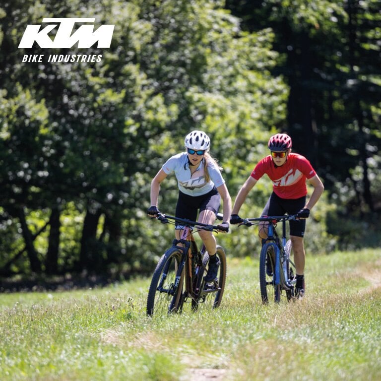 KTM Ein Mann und eine Frau, die mit ihren Fahrräder über einen Weg der über eine Wiese führt, fahren