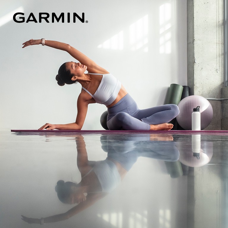 Person macht eine seitliche Dehnübung auf einer rosafarbenen Yogamatte in einem hellen Raum. Links oben ist das Logo „GARMIN“ sichtbar. Im Hintergrund stehen ein Gymnastikball, eine Trinkflasche und eine Holzbox.