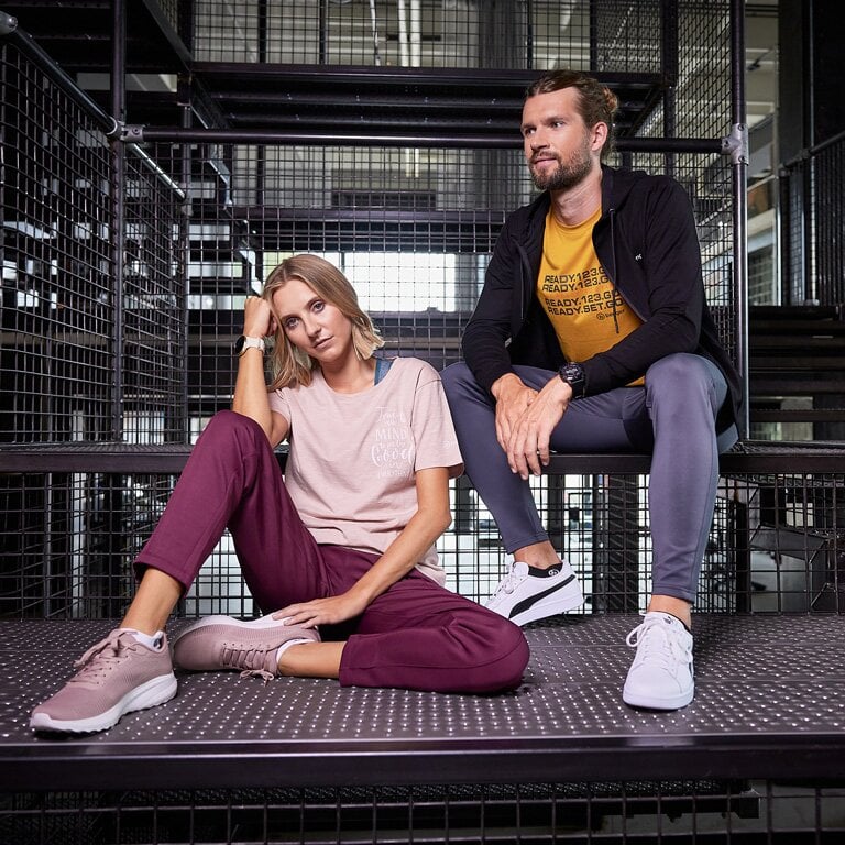 Eine Frau und ein Mann die auf eine Gitter sitzen in entspannten Outfits mit Freizeitschuhen