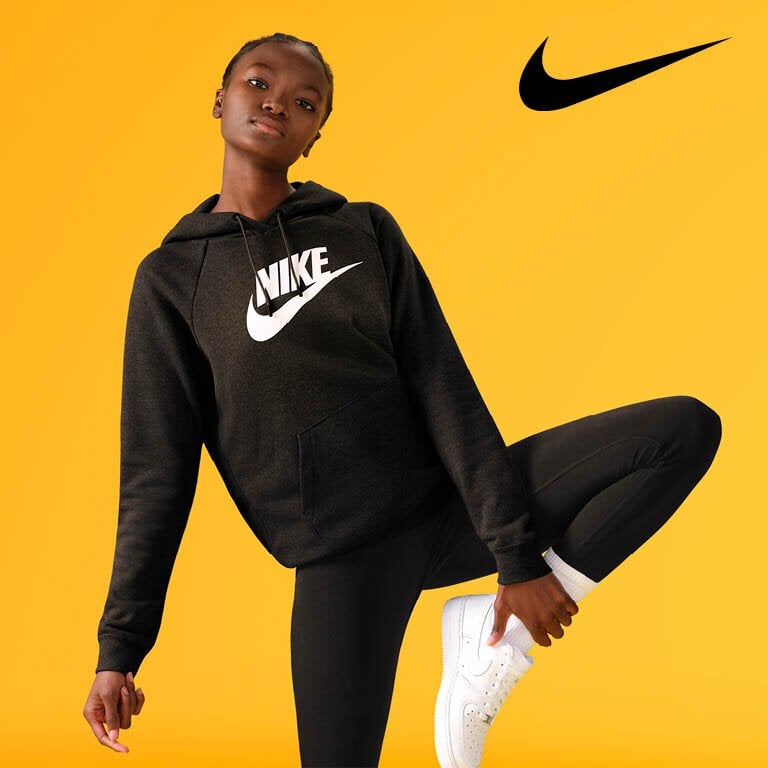 Nike EIne Frau die vor einem gelben Hintergrund steht und einen Nike Pullover und Nike Schuhe trägt
