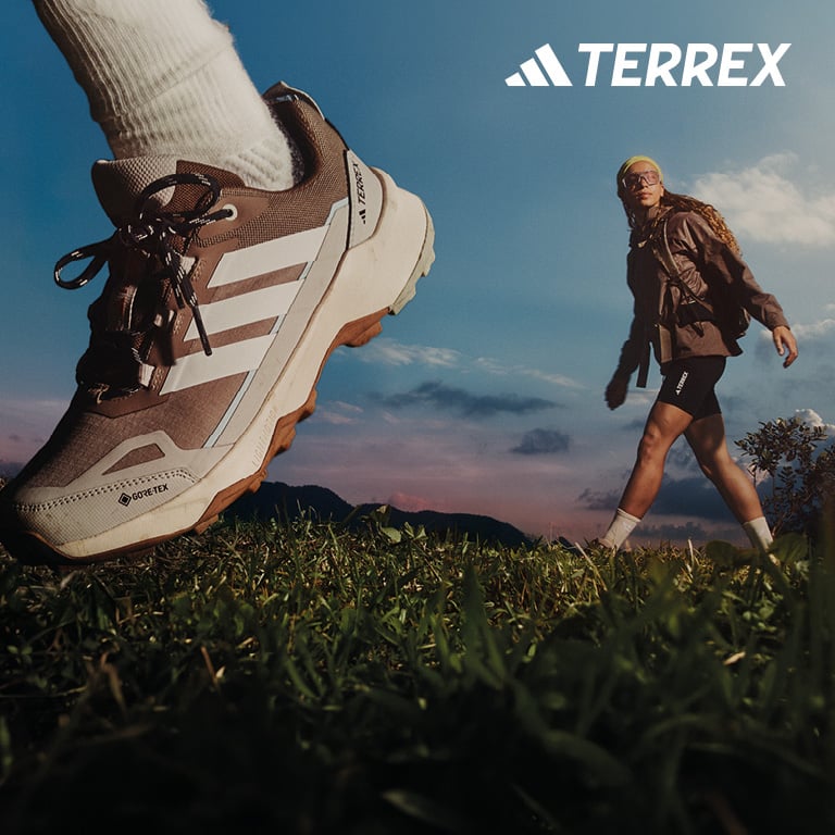 Ein großer Trail‑Schuh ist im Vordergrund zu sehen, während im Hintergrund eine Person auf einer Wiese vor einer Berglandschaft geht. Rechts oben im Bild steht das TERREX‑Logo.