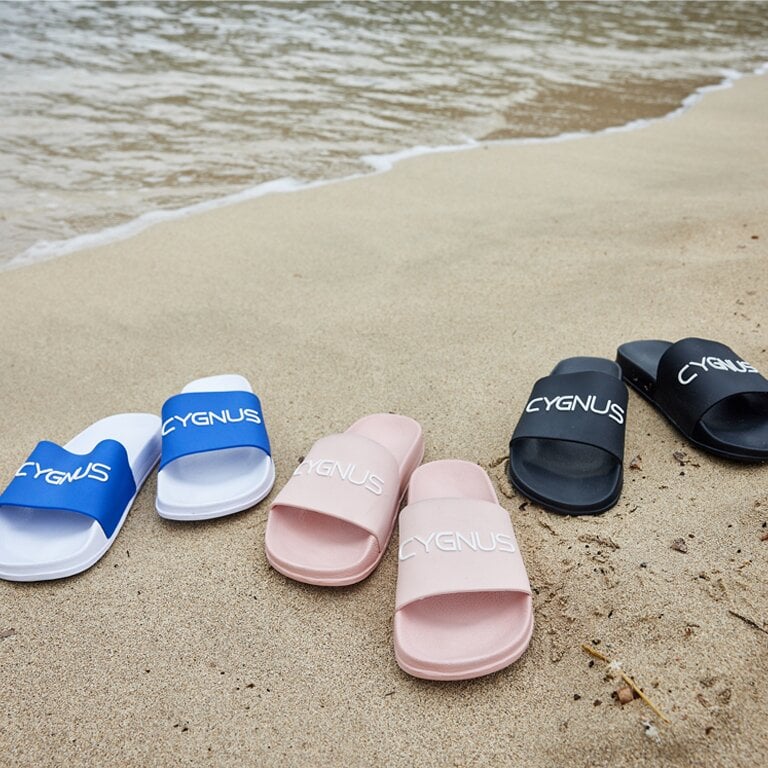 Zu sehen sind drei Cygnus Badeschlappen am Strand in blau, rosa und schwarz mit dem Cygnus Schriftzug. 
