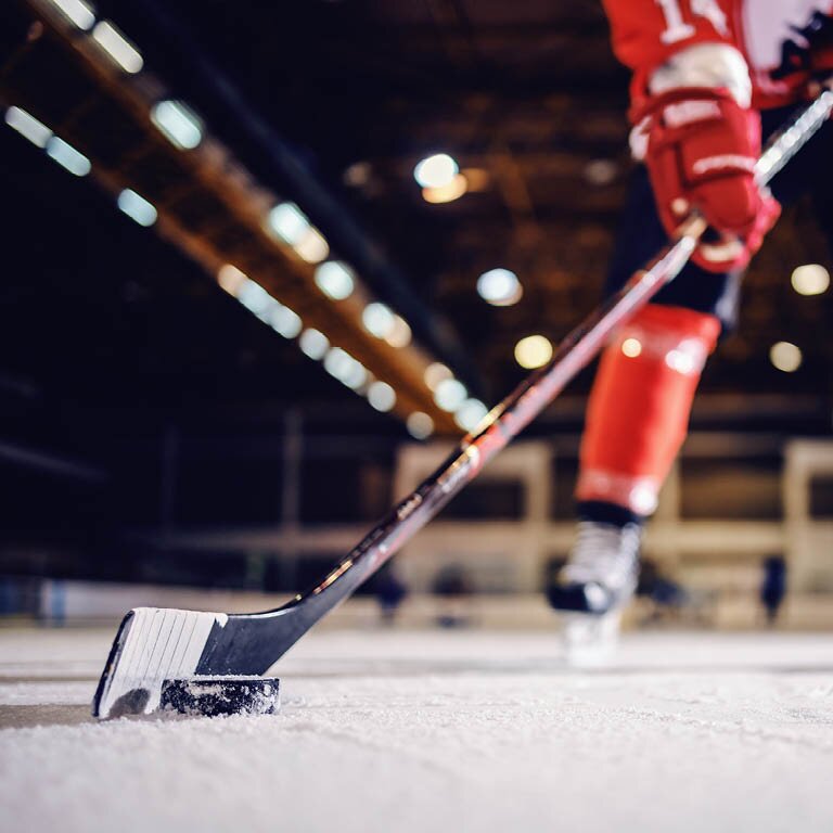 Zu sehen ist ein Eishockeyspieler am Eis mit einem Eishockeyschläger und einem Puck