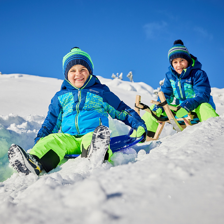 Zwei Kinder sitzen auf einer Rodel und einem Schlitten im Schnee