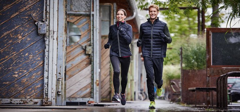 Zwei Personen joggen auf nassem Pflaster in einer städtischen Umgebung. Beide tragen dunkle Sportkleidung.
