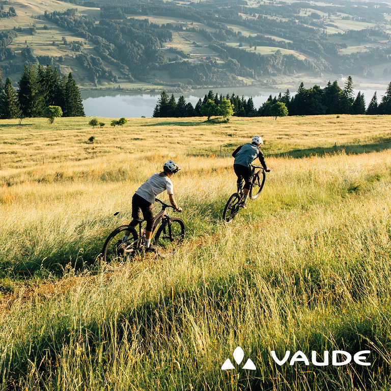Zwei Personen fahren mit Fahrrädern durch eine weitläufige Wiesenlandschaft mit Blick auf Wälder und einen See. Rechts unten im Bild steht das VAUDE‑Logo.