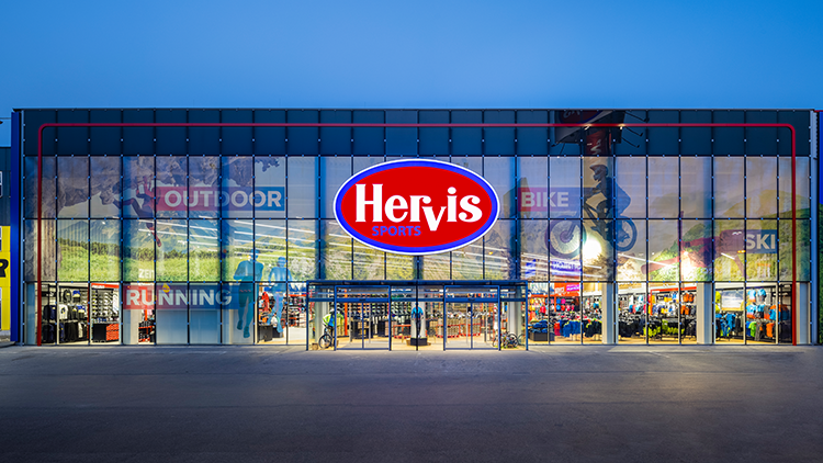 Radberater Die Außenansicht des Hervis Sports-Stores in Wien Stadlau. Die Fassade besteht aus Glaswänden, und in der Mitte befindet sich ein großes rotes und blaues Hervis Sports-Logo. Über dem Eingang sind die Wörter 'OUTDOOR', 'BIKE', 'SKI' und 'RUNNING' auf den Glaswänden zu sehen. Durch das Glas ist das Innere des Geschäfts mit verschiedenen Sportgeräten und -bekleidung sichtbar.