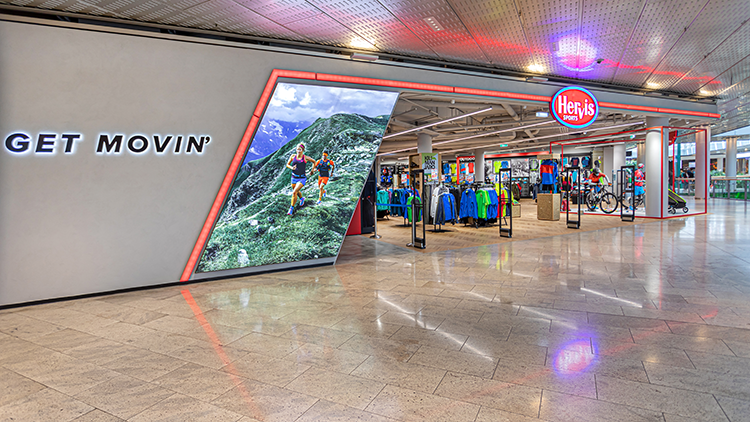 Radberater Die Außenansicht des Hervis Stores Europark Salzburg. Der Store hat einen großen Glaseingang mit dem Hervis-Logo, das gut sichtbar darüber angebracht ist. Vor dem Store befindet sich ein Parkplatz mit mehreren Büschen und einem Fußgängerüberweg, der zum Eingang führt. Im Hintergrund sind unter einem klaren blauen Himmel Berge zu sehen.