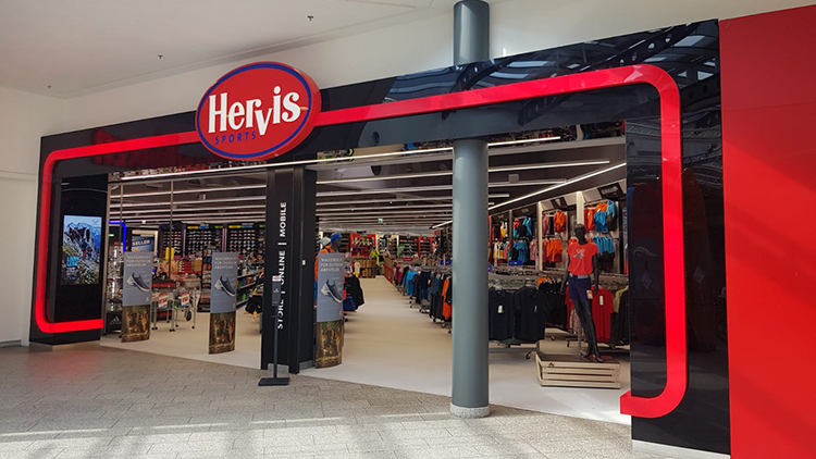 Radberater Der Eingang des Hervis Sports Stores im einem Einkaufszentrum Haid. Über dem Eingang befindet sich ein auffälliges rotes und weißes Logo mit der Aufschrift 'Hervis Sports'. Der Eingang ist von einem schwarz-roten Design umrahmt, mit Glastüren, die in den Store führen. Im Inneren sind verschiedene Sportbekleidung und -ausrüstung auf Regalen und Ständern ausgestellt.