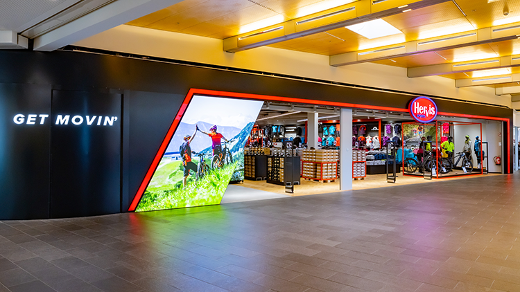 Radberater Die Fassade des Hervis Sports Stores in Graz Murpark. Der Store hat ein modernes Design mit einer schwarz-roten Außenansicht. Auf der linken Seite befindet sich ein beleuchtetes Schild mit der Aufschrift 'GET MOVIN''. Der Eingang besteht aus großen Glasfenstern, die verschiedene Sportgeräte und -bekleidung im Inneren des Stores zeigen. Das Innere des Stores ist gut beleuchtet und präsentiert Artikel wie Fahrräder, Kleidung und andere Sportartikel.